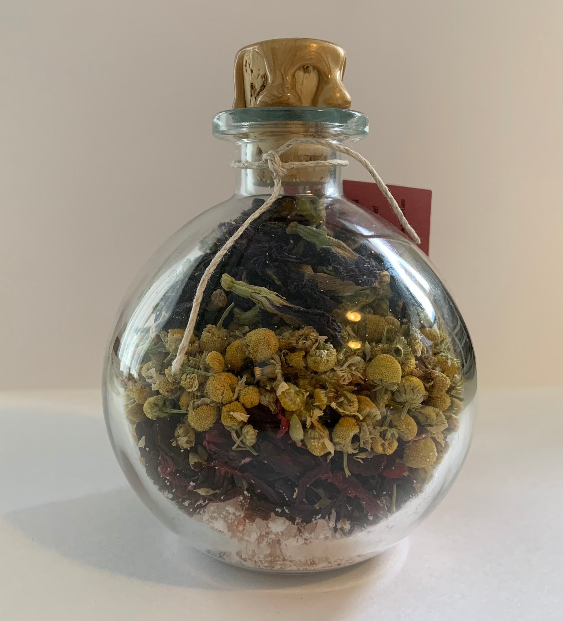 Bain floral Flower Moon sur arrière-plan blanc. Focus sur les étages d'ingrédients : Sel d'Himalaya et Poudre de lait de Coco, pétales d'Hibiscus, bourgeons de Camomille, pétales de fleur de Pois Papillon.
