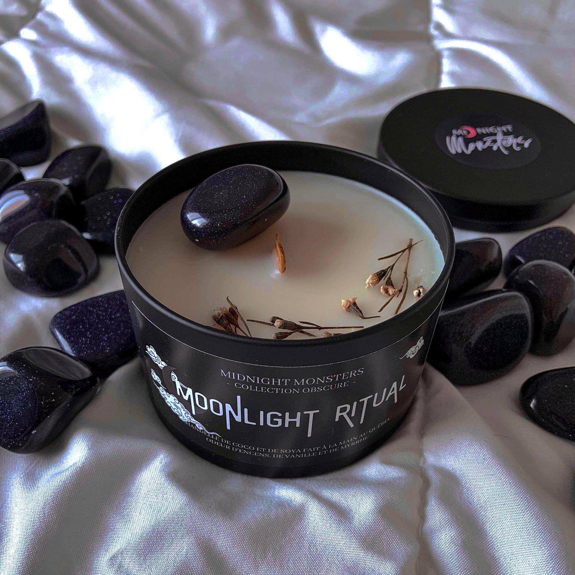 Chandelle Moonlight Ritual, couvercle retiré, entourée de pierre de Goldstone bleue. Focus sur la décoration de la chandelle, fleurs de Wax séchée et une Goldstone bleue.